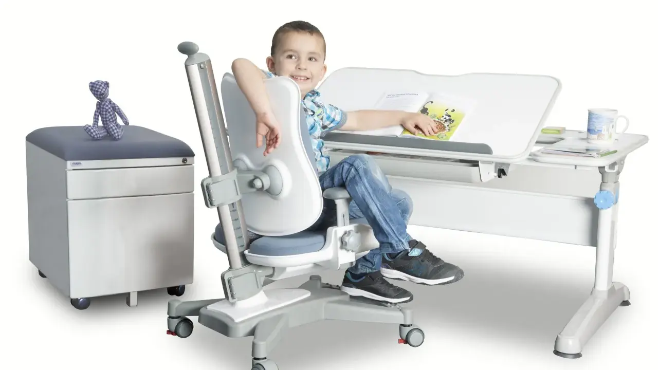 Uśmiechnięty chłopiec siedzi przy biurku na ergonomicznym krześle, czytając książkę.