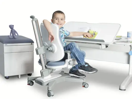 Uśmiechnięty chłopiec siedzi przy biurku na ergonomicznym krześle, czytając książkę.