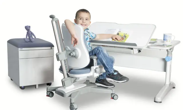 Uśmiechnięty chłopiec siedzi przy biurku na ergonomicznym krześle, czytając książkę.